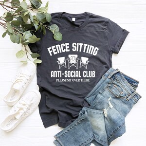May include: A dark grey t-shirt with white text that reads "Fence Sitting Anti-Social Club Please Sit Over There". The text is surrounded by a graphic of three chairs with a heart in the middle.