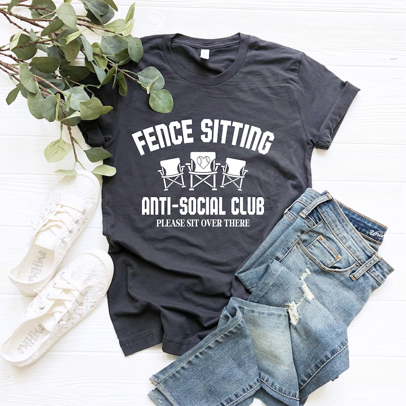 May include: A dark grey t-shirt with white text that reads "Fence Sitting Anti-Social Club Please Sit Over There". The text is surrounded by a graphic of three chairs with a heart in the middle.