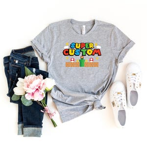 May include: A gray t-shirt with the words "SUPER CUSTOM" in a colorful video game-style font. The shirt features a brick platform, mushrooms, and a green pipe design. The shirt is paired with blue jeans, white sneakers, and a bouquet of pink and white flowers.