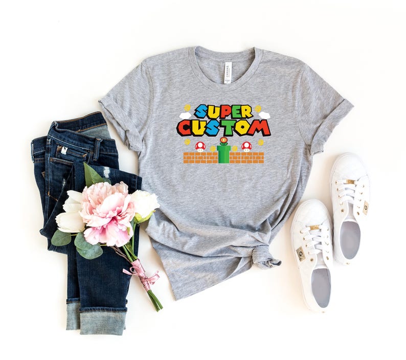 May include: A gray t-shirt with the words "SUPER CUSTOM" in a colorful video game-style font. The shirt features a brick platform, mushrooms, and a green pipe design. The shirt is paired with blue jeans, white sneakers, and a bouquet of pink and white flowers.