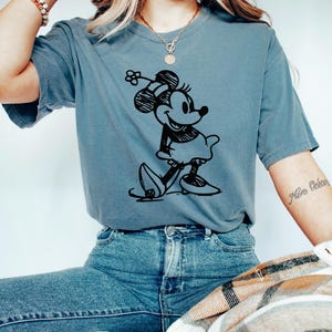 May include: A blue t-shirt featuring a black graphic of Minnie Mouse. The cartoon character is depicted in a classic pose, wearing a dress and hat. The shirt is paired with blue jeans.