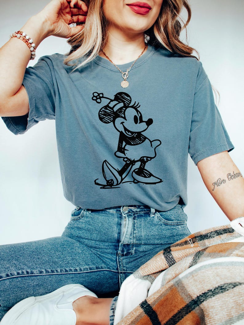May include: A blue t-shirt featuring a black graphic of Minnie Mouse. The cartoon character is depicted in a classic pose, wearing a dress and hat. The shirt is paired with blue jeans.
