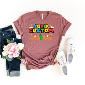 May include: A mauve t-shirt with the words "SUPER CUSTOM" in a colorful, video game-inspired font. The design includes pixelated elements like coins, mushrooms, and a green pipe. The shirt is paired with blue jeans, white sneakers, and a bouquet of pink flowers.