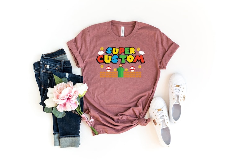 May include: A mauve t-shirt with the words "SUPER CUSTOM" in a colorful, video game-inspired font. The design includes pixelated elements like coins, mushrooms, and a green pipe. The shirt is paired with blue jeans, white sneakers, and a bouquet of pink flowers.