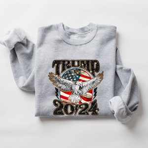 May include: Gray sweatshirt with a graphic of an eagle flying in front of a circle with an American flag and the text "TRUMP 2024".