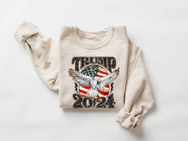 May include: A beige sweatshirt with a black graphic print of an eagle flying over an American flag with the text "TRUMP 2024".