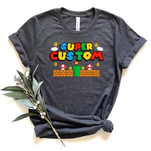May include: Dark gray t-shirt featuring the text "SUPER CUSTOM" in a colorful, video game-inspired font. The design includes pixelated mushrooms, coins, and a green pipe, reminiscent of a classic video game.