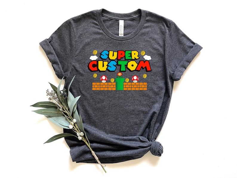 May include: Dark gray t-shirt featuring the text "SUPER CUSTOM" in a colorful, video game-inspired font. The design includes pixelated mushrooms, coins, and a green pipe, reminiscent of a classic video game.