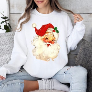 May include: White sweatshirt with a vintage illustration of Santa Claus's face. The illustration features a red hat with white trim and a white beard.