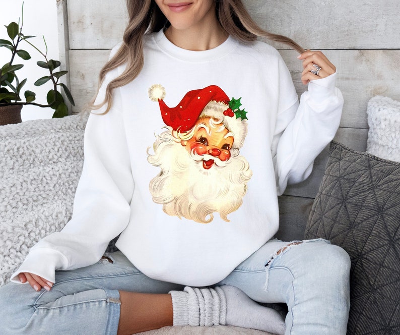 May include: White sweatshirt with a vintage illustration of Santa Claus's face. The illustration features a red hat with white trim and a white beard.