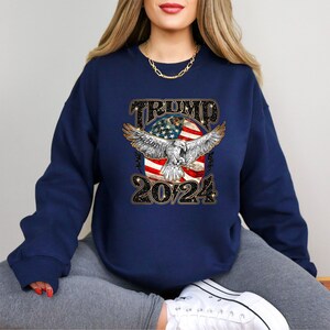 May include: A navy blue sweatshirt with a graphic of an eagle with wings spread, a red, white, and blue American flag, and the text "TRUMP 2024".
