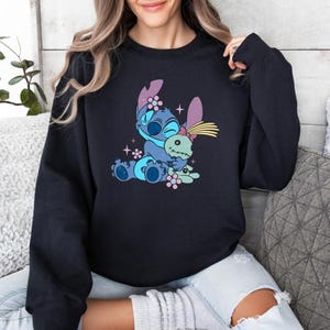 K&ouml;nnte beinhalten: Schwarzes Sweatshirt mit einem Cartoon-Motiv von Stitch, der eine Puppe umarmt. Die Grafik ist in Blau, Rosa und Gelb gehalten. Das Sweatshirt hat einen Rundhalsausschnitt.