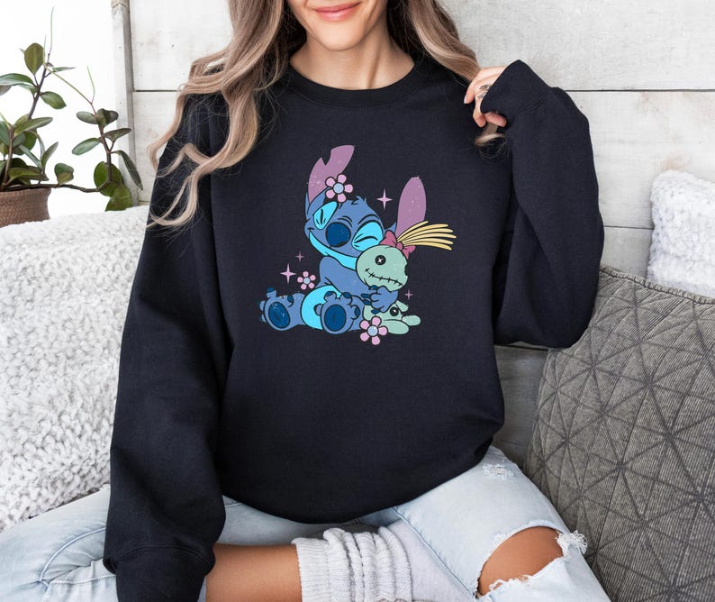 K&ouml;nnte beinhalten: Schwarzes Sweatshirt mit einem Cartoon-Motiv von Stitch, der eine Puppe umarmt. Die Grafik ist in Blau, Rosa und Gelb gehalten. Das Sweatshirt hat einen Rundhalsausschnitt.