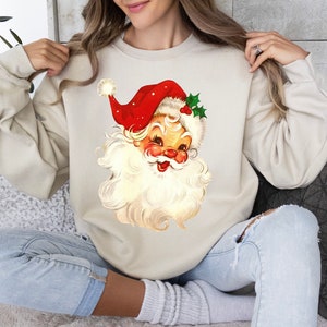 May include: A cream-colored sweatshirt with a vintage-style illustration of Santa Claus on the front. Santa is wearing a red hat with white trim and a white beard. He is smiling and has a red nose.