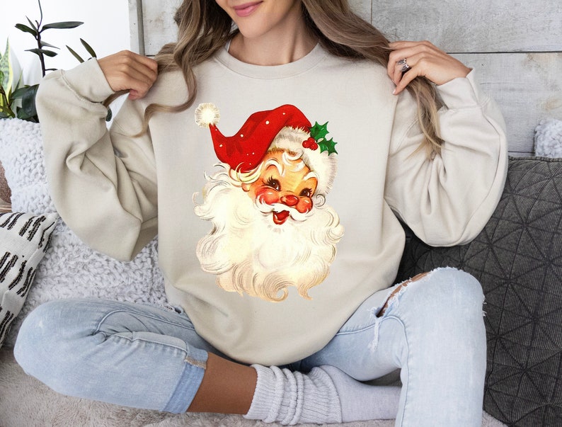 May include: A cream-colored sweatshirt with a vintage-style illustration of Santa Claus on the front. Santa is wearing a red hat with white trim and a white beard. He is smiling and has a red nose.