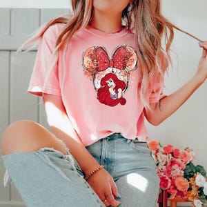 May include: A pink t-shirt with a graphic of Ariel from The Little Mermaid in a Mickey Mouse ear headband. The headband is red with white polka dots and the graphic is surrounded by a watercolor-like design.