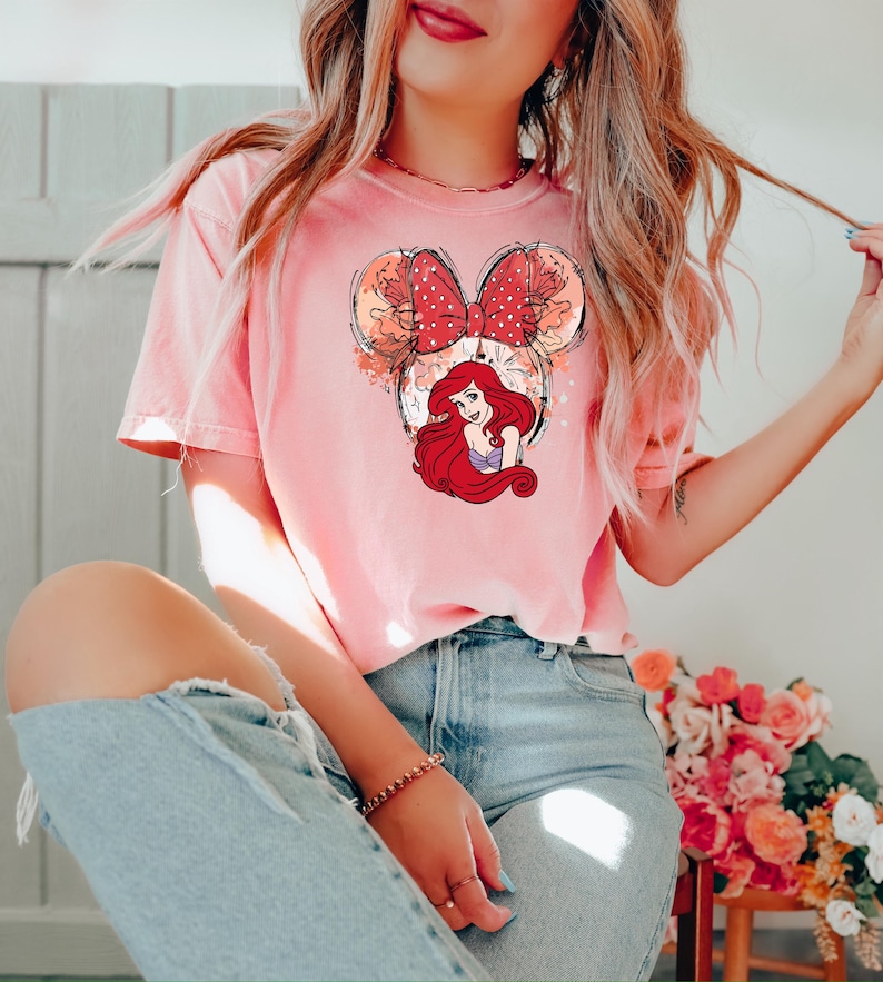 May include: A pink t-shirt with a graphic of Ariel from The Little Mermaid in a Mickey Mouse ear headband. The headband is red with white polka dots and the graphic is surrounded by a watercolor-like design.