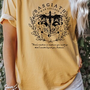 Peut inclure: Un t-shirt jaune avec un motif graphique noir repr&eacute;sentant un dragon, une couronne et le texte "BASGIATH WAR COLLEGE". Le texte sous le graphique indique "There's nowhere in existence you could go that I wouldn't find you, Violence."