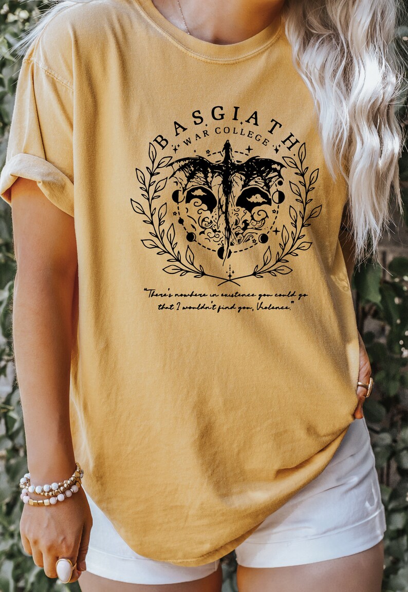Peut inclure: Un t-shirt jaune avec un motif graphique noir repr&eacute;sentant un dragon, une couronne et le texte "BASGIATH WAR COLLEGE". Le texte sous le graphique indique "There's nowhere in existence you could go that I wouldn't find you, Violence."