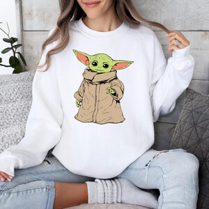 May include: White sweatshirt with a green and brown cartoon character wearing a brown robe. The character has large ears and is holding its hand up.