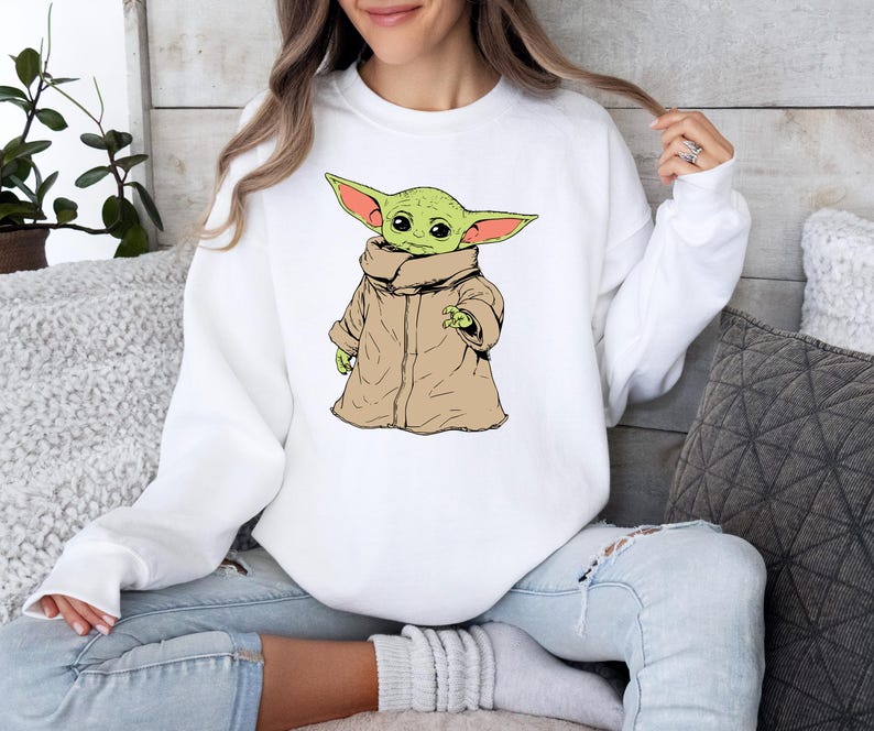May include: White sweatshirt with a green and brown cartoon character wearing a brown robe. The character has large ears and is holding its hand up.