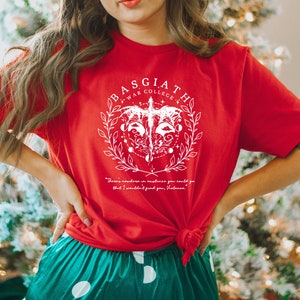 May include: Red t-shirt with a white graphic design featuring a bird with a crown and the text "Basgiath War College". The text "There's nowhere in existence you could go that I wouldn't find you, Volence." is printed below the graphic.