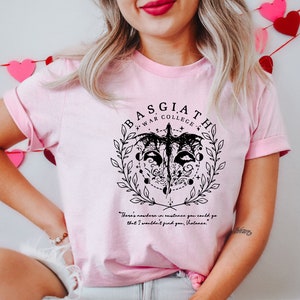 May include: Pink t-shirt with a black graphic design featuring a stylized bird with a circle around it. The text "BASGIATH WAR COLLEGE" is above the bird. The quote "There's nowhere in existence you could go that I wouldn't find you, Volana." is below the bird.