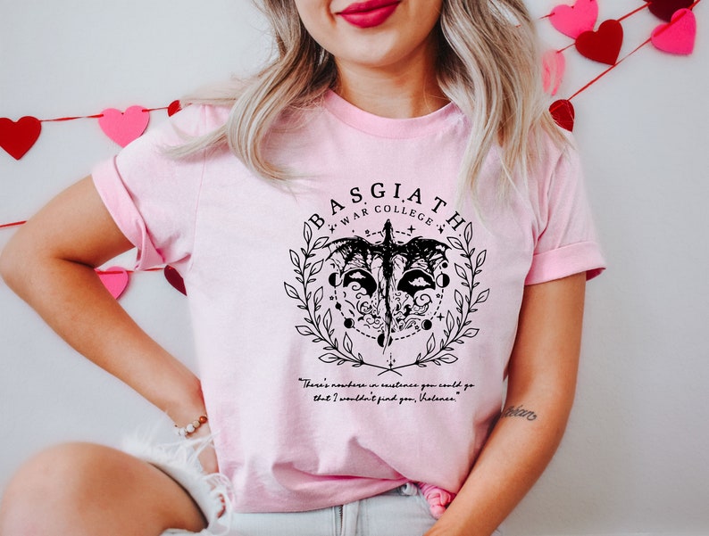 May include: Pink t-shirt with a black graphic design featuring a stylized bird with a circle around it. The text "BASGIATH WAR COLLEGE" is above the bird. The quote "There's nowhere in existence you could go that I wouldn't find you, Volana." is below the bird.