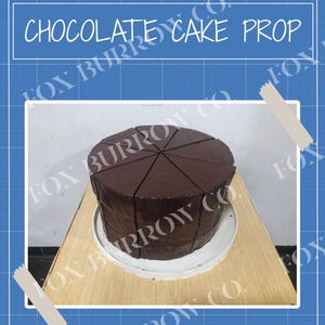 May include: A chocolate cake prop, cut into slices, sits on a white plate atop a wooden surface. The image is framed by a blue background with the text "CHOCOLATE CAKE PROP" and the Fox Burrow Co. logo.