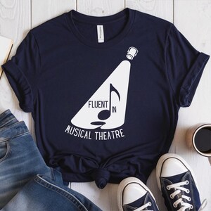 Op de afbeelding: Donkerblauw t-shirt met een witte afbeelding van een schijnwerper die op een noot schijnt. De tekst "Fluent in Musical Theatre" staat onder de afbeelding.