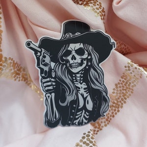 May include: Black and white sticker of a skeleton woman wearing a cowboy hat and holding a gun. The sticker is on a pink fabric background with gold glitter.