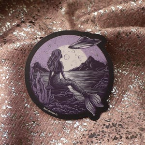 May include: A round sticker featuring a mermaid sitting on a rock, with a UFO hovering above. The design is in shades of purple and black, with a full moon in the background. The text "Sea Orange FL" is at the bottom.