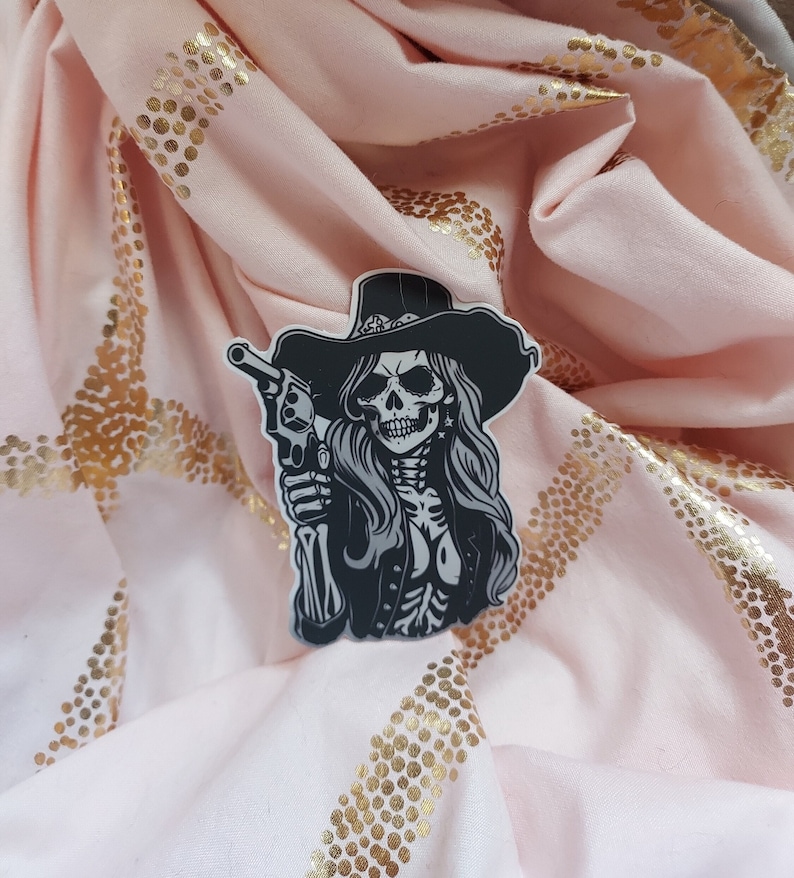 May include: Black and white sticker of a skeleton woman wearing a cowboy hat and holding a gun. The sticker is on a pink fabric background with gold glitter.