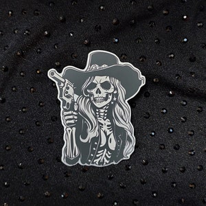 May include: A black and white sticker of a skull wearing a cowboy hat and holding a gun. The skull has a detailed design with a long, flowing mane of hair.