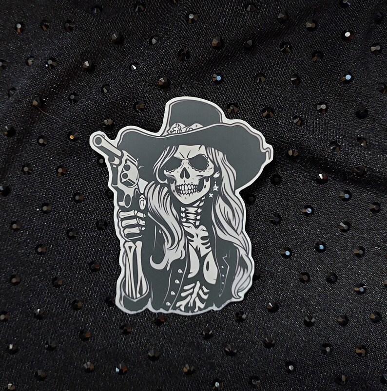May include: A black and white sticker of a skull wearing a cowboy hat and holding a gun. The skull has a detailed design with a long, flowing mane of hair.
