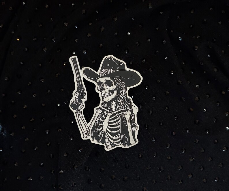 Peut inclure: Un autocollant noir et blanc d'un squelette portant un chapeau de cowboy et tenant un pistolet. Le squelette porte un gilet et a les cheveux longs.