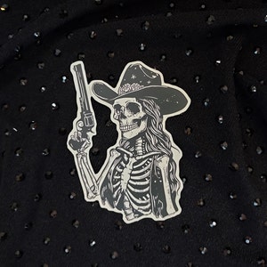Peut inclure: Autocollant noir et blanc d'un squelette portant un chapeau de cowboy et tenant un pistolet. Le squelette a les cheveux longs et porte un gilet.