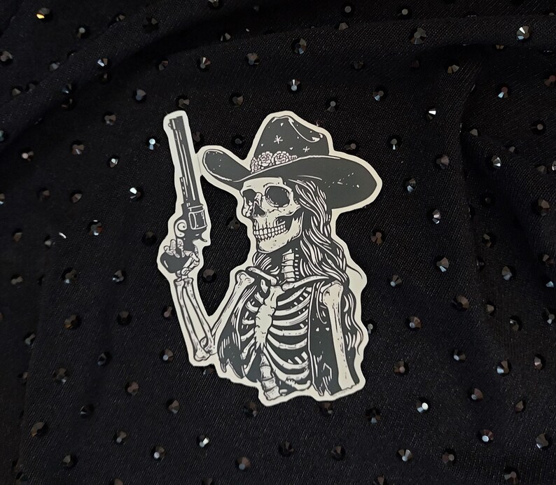 Peut inclure: Autocollant noir et blanc d'un squelette portant un chapeau de cowboy et tenant un pistolet. Le squelette a les cheveux longs et porte un gilet.