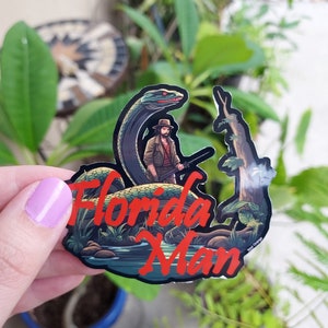 May include: A sticker featuring a cartoon illustration of a man with a snake, with the words "Florida Man" in red. The man is wearing a hat and holding a weapon. The background includes a river and trees.
