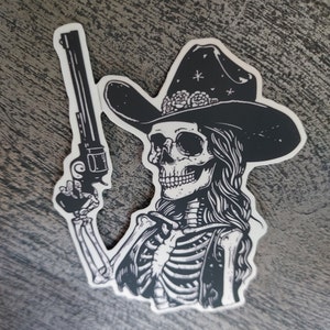 Peut inclure: Illustration en noir et blanc d'un squelette portant un chapeau de cowboy avec des fleurs et tenant un pistolet.