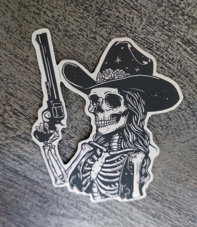 Peut inclure: Illustration en noir et blanc d'un squelette portant un chapeau de cowboy avec des fleurs et tenant un pistolet.