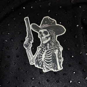 Peut inclure: Un autocollant noir et blanc d'un squelette portant un chapeau de cowboy et tenant un pistolet. L'autocollant est sur un fond noir.