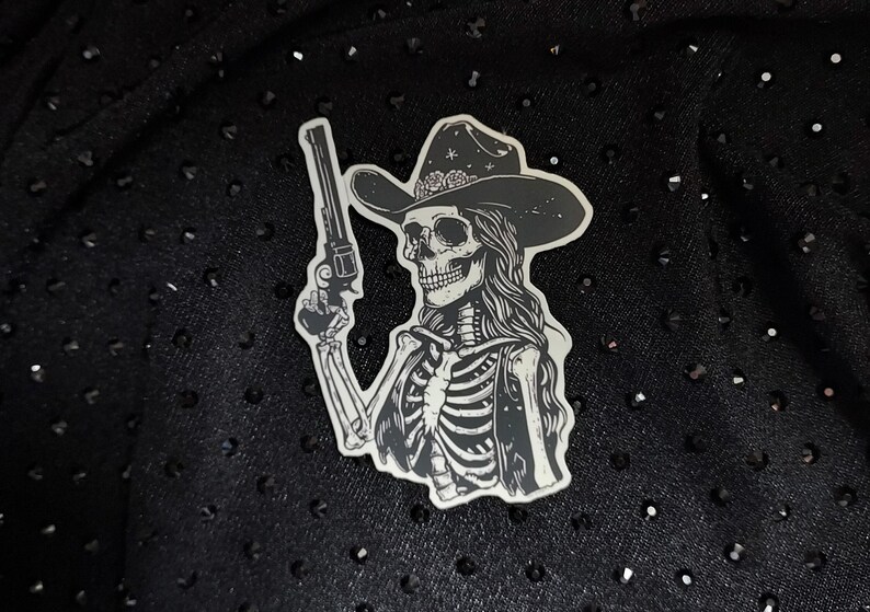 Peut inclure: Un autocollant noir et blanc d'un squelette portant un chapeau de cowboy et tenant un pistolet. L'autocollant est sur un fond noir.