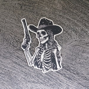 Peut inclure: Illustration en noir et blanc d'un squelette portant un chapeau de cowboy et tenant un pistolet. Le squelette porte un gilet et a les cheveux longs.