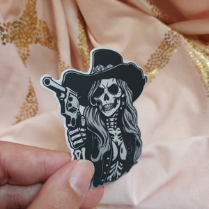 May include: Black and white sticker of a skull wearing a cowboy hat and holding a gun. The skull has long hair and is wearing a jacket.