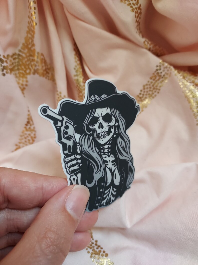 May include: Black and white sticker of a skull wearing a cowboy hat and holding a gun. The skull has long hair and is wearing a jacket.