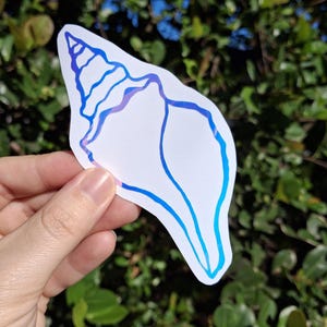Könnte beinhalten: Ein weißer Muschelschalen-Aufkleber mit blau-violettem Farbverlauf. Der Aufkleber wird vor dem Hintergrund von grünem Laub und blauem Himmel gehalten. Das Design zeigt detaillierte Linien.