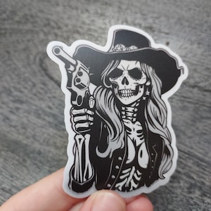 May include: Black and white sticker of a skeleton woman wearing a cowboy hat and holding a gun.
