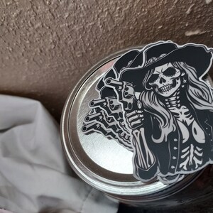 May include: Black and white sticker of a skull wearing a cowboy hat and holding two pistols. The skull has a bandana around its head and is wearing a jacket. The sticker is on a silver jar lid.