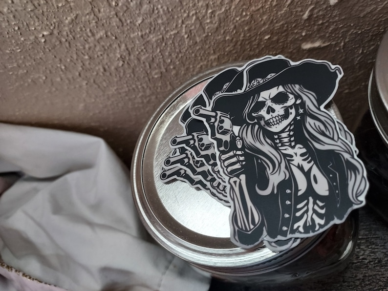 May include: Black and white sticker of a skull wearing a cowboy hat and holding two pistols. The skull has a bandana around its head and is wearing a jacket. The sticker is on a silver jar lid.